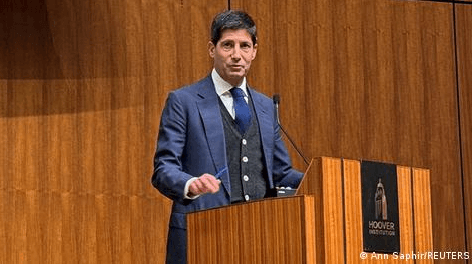 Kevin Warsh öltönyös férfi egy pulpitusnál áll és beszédet tart a Hoover Institution rendezvényén. A kép jobb sarkában az Ann Saphir/REUTERS felirat látható.
